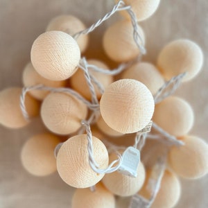 May include: A string of cream-colored cotton ball lights. The lights are connected by a white wire and have a white plug.