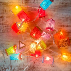 K&ouml;nnte beinhalten: Eine Lichterkette mit bunten Papierlaternen und warmwei&szlig;en Lichtern. Die Laternen haben verschiedene Formen, darunter Quadrate, Rechtecke und Dreiecke. Die Lichter sind an einem wei&szlig;en Kabel befestigt und h&auml;ngen an einer Holzoberfl&auml;che.