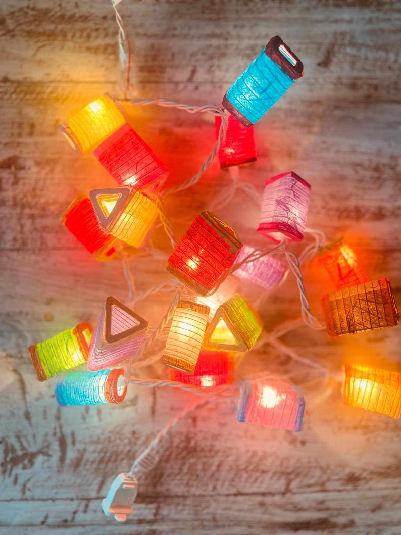 K&ouml;nnte beinhalten: Eine Lichterkette mit bunten Papierlaternen und warmwei&szlig;en Lichtern. Die Laternen haben verschiedene Formen, darunter Quadrate, Rechtecke und Dreiecke. Die Lichter sind an einem wei&szlig;en Kabel befestigt und h&auml;ngen an einer Holzoberfl&auml;che.
