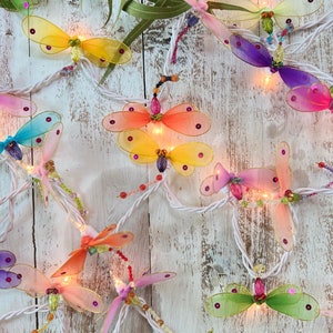 Puede incluir: Una cadena de luces de hadas con bombillas en forma de libélula en varios colores, incluyendo rosa, amarillo, verde y azul. Las luces están enhebradas en un cable blanco y están iluminadas, creando un brillo cálido y acogedor.