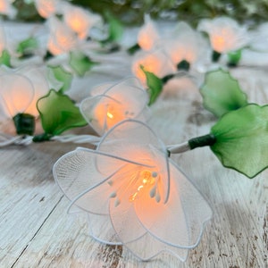 May include: A decorative string of white fabric flower lights with green leaves. Each flower contains a warm, glowing light. The string is on a white cable, ideal for adding a soft, decorative touch to any space.