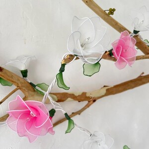 May include: A string of fairy lights with white and pink fabric flowers. The lights are strung on a wire with green leaves. The flowers are made of a sheer fabric and have a delicate, feminine look.