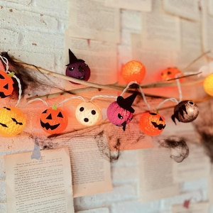 May include: A string of Halloween-themed lights with orange jack-o'-lanterns, purple witch hats, and other decorative spheres. The lights are strung on a branch against a backdrop of aged book pages and a white brick wall.