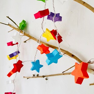 May include: A string of colorful star-shaped lights, in shades of red, orange, yellow, green, blue, and purple, are strung along a white wire. The lights are attached to a natural branch against a white background.