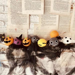 May include: A collection of handmade Halloween-themed plush toys, including a black cat, pumpkins, spiders, and ghosts. The toys are arranged on a wooden stick against a backdrop of vintage book pages and black spiderweb decor.