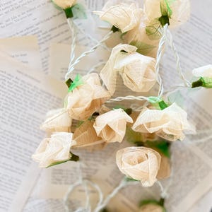 Pode incluir: Uma corda de luzes com delicadas flores de papel em forma de rosa de cor creme. As luzes são enfiadas em um fio branco com folhas verdes intercaladas entre as flores.