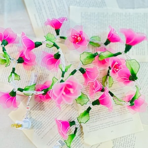 May include: A string of pink flower fairy lights with green leaves. The lights are strung on a white wire and have a white plug.