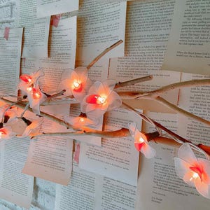 May include: A string of decorative fairy lights with white flower-shaped shades and warm orange lights. The lights are arranged on a branch against a backdrop of vintage book pages and a white brick wall.