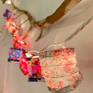 Puede incluir: Una cadena de luces blancas con farolillos de papel. Los farolillos están decorados con un patrón floral en tonos rosa, blanco y verde. Las luces están colgadas de una rama.