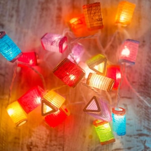 K&ouml;nnte beinhalten: Eine Lichterkette mit bunten Papierlaternen, die von innen beleuchtet werden. Die Laternen haben verschiedene Formen, darunter Quadrate, Dreiecke und Rechtecke. Die Lichter sind eingeschaltet und leuchten, wodurch eine warme und festliche Atmosph&auml;re entsteht.