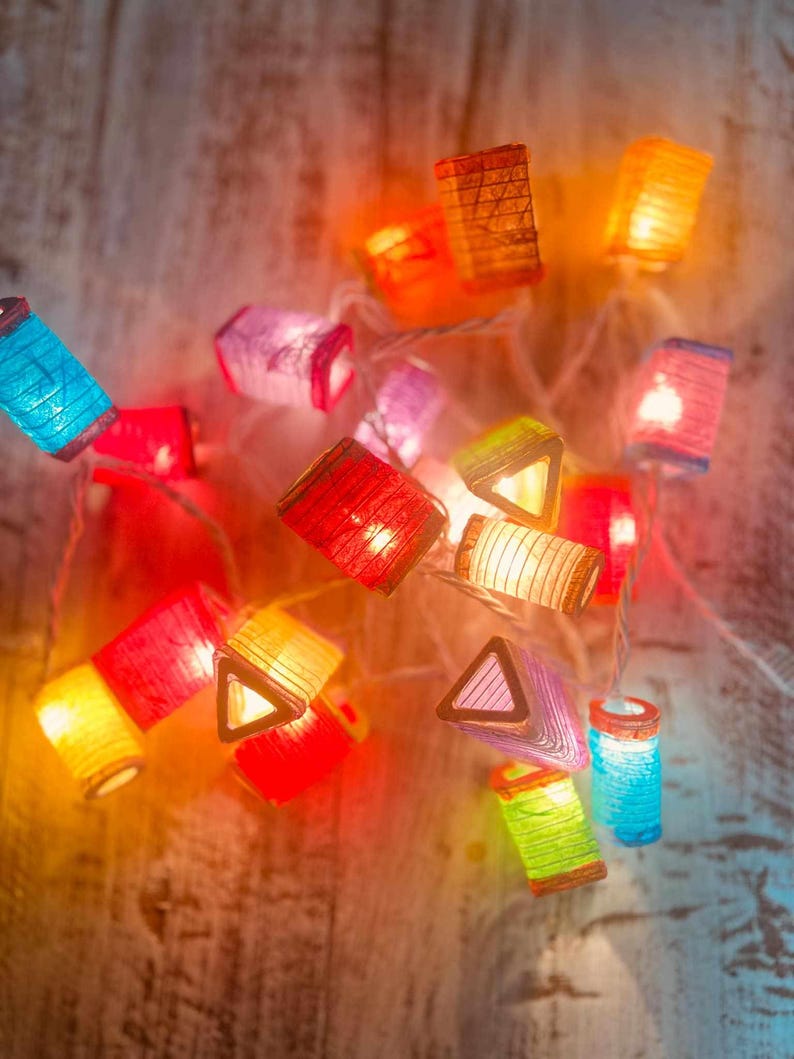 K&ouml;nnte beinhalten: Eine Lichterkette mit bunten Papierlaternen, die von innen beleuchtet werden. Die Laternen haben verschiedene Formen, darunter Quadrate, Dreiecke und Rechtecke. Die Lichter sind eingeschaltet und leuchten, wodurch eine warme und festliche Atmosph&auml;re entsteht.