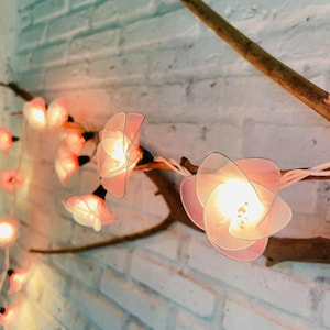 Op de afbeelding: Een reeks decoratieve lampjes in de vorm van roze bloemen, bevestigd aan een bruine tak. De lampjes zijn aan, waardoor een warme gloed ontstaat tegen een witte bakstenen muur. De bloemen hebben een delicaat ontwerp.