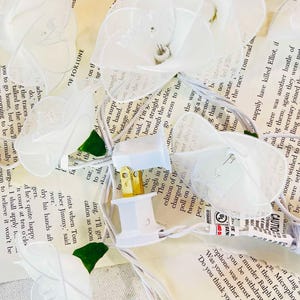 May include: White string lights with clear plastic flower-shaped bulbs. The lights are plugged into an electrical outlet and have a white cord.