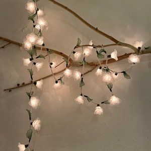 Puede incluir: Luces de cadena de flores blancas con hojas verdes envueltas alrededor de una rama. Las luces están conectadas con un cable transparente y emiten un brillo blanco cálido.