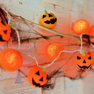 May include: A string of orange pumpkin-shaped lights with black cut-out faces and green stems. The lights are strung on a white wire and are displayed against a backdrop of aged paper and a light-colored wall. Halloween decor.