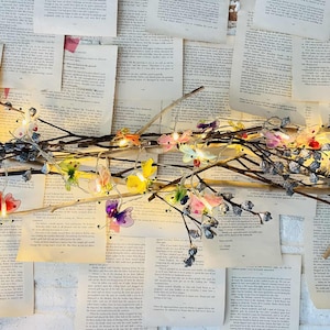 Pode incluir: Uma guirlanda de luzes decorativa com ornamentos em forma de borboleta coloridos e pequenas luzes, fixada a um galho marrom. O galho é colocado sobre um fundo de páginas de livros antigos, criando uma estética caprichosa e inspirada na natureza.