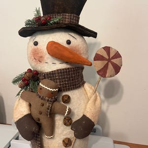 May include: A fabric snowman figurine wearing a dark brown top hat with pine and berry accents, a brown plaid scarf, and metal jingle bell buttons. It holds a brown gingerbread person doll and a round red and beige striped lollipop on a stick.
