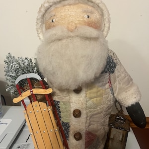 May include: A Santa Claus figurine wearing a coat made of quilted fabric patches in cream, blue, and red tones. The figure has a white faux fur beard and hat trim, holding a miniature red and wood sled with a small tree and a dark metal lantern.