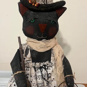 May include: A black fabric cat doll wearing a tall, dark, weathered witch hat with a '31' label, a beige cheesecloth scarf, and a white apron printed with black text. The cat holds a small broom and a brown cauldron labeled 'Witch rides $.10'.