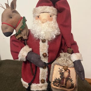 May include: A fabric Santa Claus figurine wearing a deep red coat and hat with cream-colored faux fur trim and small metal bells. Santa holds a brown fabric hobby horse with a green garland and a burlap sack featuring a printed image of Santa.