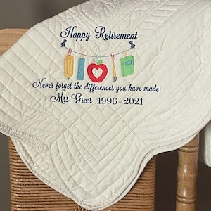 May include: A white quilted blanket with the text "Happy Retirement" embroidered in blue. The blanket also features a string of school supplies hanging from clothespins with the text "Never forget the differences you have made! Mrs. Greer 1996-2021" embroidered below.