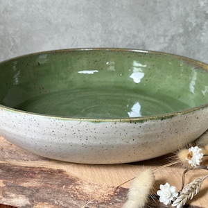 May include: A large ceramic bowl with a speckled white exterior and a glossy green interior. The bowl has a wide, shallow design, ideal for serving salads or displaying decorative items. The rim has a subtle, natural colour.