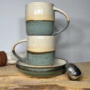 May include: Two stacked ceramic mugs with a speckled cream top, yellow band, and teal blue base. A matching saucer and a stainless steel tea infuser are also present. The mugs have curved handles and sit on a wooden surface.