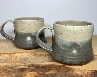 May include: Two handmade ceramic mugs with a speckled glaze. The mugs are a light beige color on top and a dark gray color on the bottom. The mugs have a unique, slightly curved handle.
