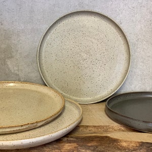 May include: Three ceramic plates, two in a light beige colour with a speckled finish and one in a dark grey colour with a smooth finish.