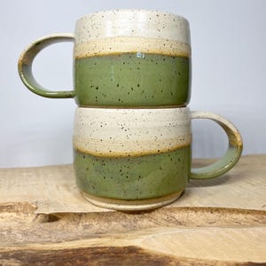 May include: Two ceramic mugs stacked on top of each other. The mugs are green, white and brown with a speckled glaze. The mugs have a rounded shape and a handle.