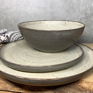 May include: A ceramic dinner set comprising a large bowl and two plates. The set has a speckled, off-white interior and a grey exterior with a dark brown rim. The pieces are arranged on a wooden surface.