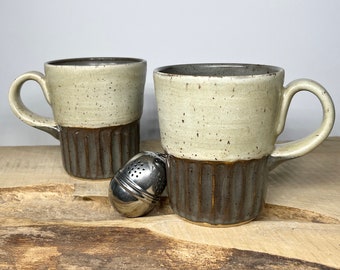 May include: Two ceramic mugs with a speckled glaze in shades of beige and brown. The mugs have a wide, rounded shape and a handle on one side. A small metal tea infuser is in the foreground.