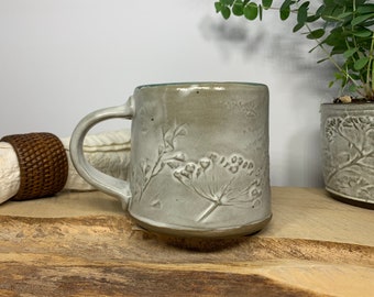 May include: A gray ceramic mug with a floral design. The mug has a handle and is sitting on a wooden surface.