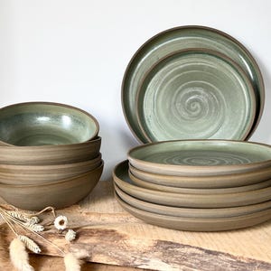 Peut inclure: Un ensemble de vaisselle en céramique comprenant une pile de trois bols et une pile de six assiettes. Les pièces présentent un extérieur brun mat et un intérieur vert sauge avec un motif en spirale visible sur la surface des assiettes et des bols.