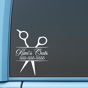 May include: A white vinyl decal applied to a dark window, featuring a graphic of a pair of scissors. The text Kim's Cuts is written in a cursive script, followed by the phone number 555-555-5555, all centered within the scissor design.