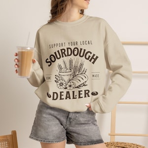 May include: A beige crewneck sweatshirt featuring dark brown text that reads SUPPORT YOUR LOCAL SOURDOUGH DEALER, along with a graphic illustration of a loaf of bread, wheat stalks, and flowers. The sweatshirt has long sleeves and a ribbed collar.