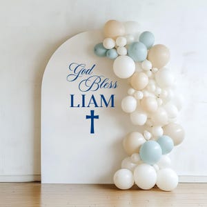 May include: A white arch-shaped backdrop with the words "God Bless LIAM" and a cross in navy blue. A balloon garland in white, light blue, and beige adorns the right side, creating a festive display.