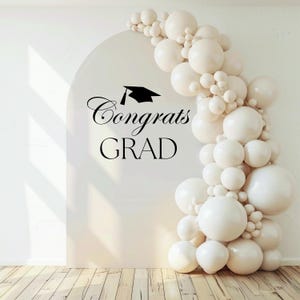 May include: A graduation party backdrop featuring a white arch with the words "Congrats GRAD" in black script and a mortarboard icon. A garland of white balloons decorates the right side of the arch, creating a festive atmosphere.