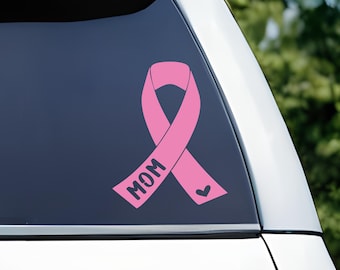 May include: A pink awareness ribbon decal applied to a vehicle rear window. The ribbon features the word MOM printed in black capital letters along the left side and a small black heart icon positioned on the right tail of the ribbon design.