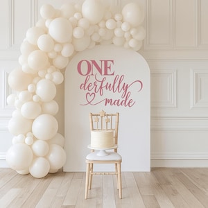 May include: A celebratory scene with a white cake on a small chair, adorned with star-shaped toppers. A large arch with the text "ONE derfully made" in pink is behind the chair. A balloon arch in cream color frames the scene.