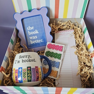 May include: A gift box with a striped lid containing a lavender wavy frame with the text 'the book was better.', a white mug with a blue handle and floral book spine print reading 'Sorry, I'm booked.', a library card notepad, and scalloped stickers.