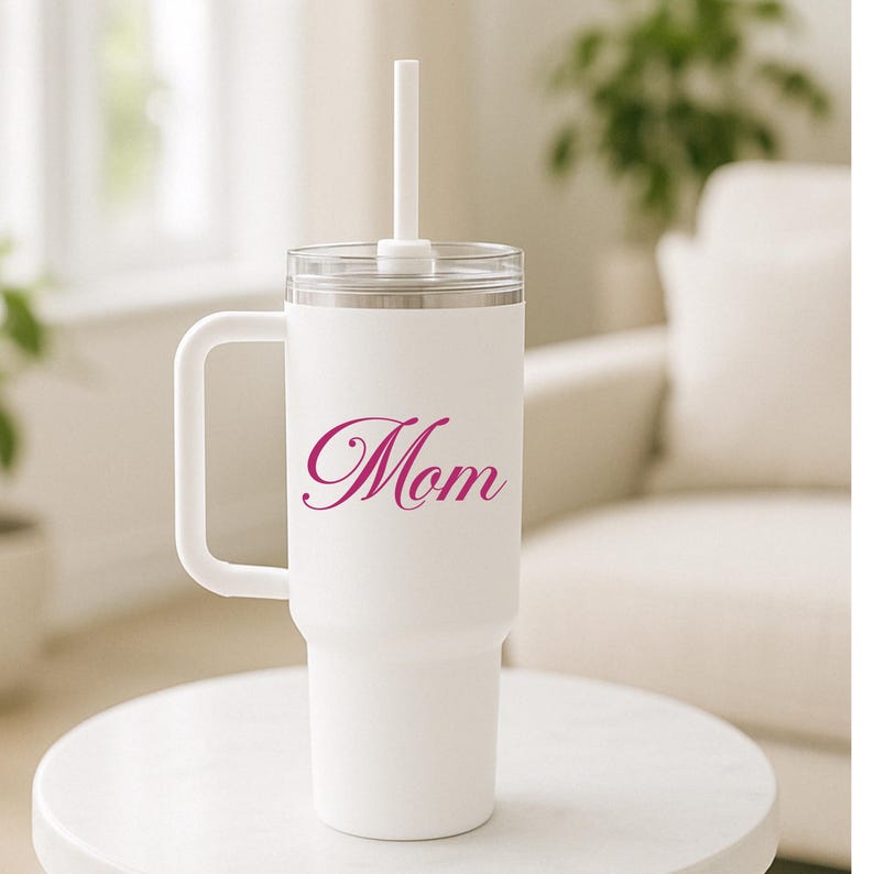 May include: A white stainless steel tumbler with a handle, a clear lid, and a white straw. The word Mom is printed in a magenta cursive script on the front of the tumbler. The cup has a tapered base and a smooth matte finish on the exterior surface.