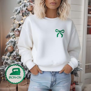 May include: A white long-sleeve sweatshirt featuring a small green bow graphic on the upper left chest. The garment has a crew neckline, ribbed cuffs, and a ribbed hem. A circular green badge with an iron icon and the text IRON-ON is visible.