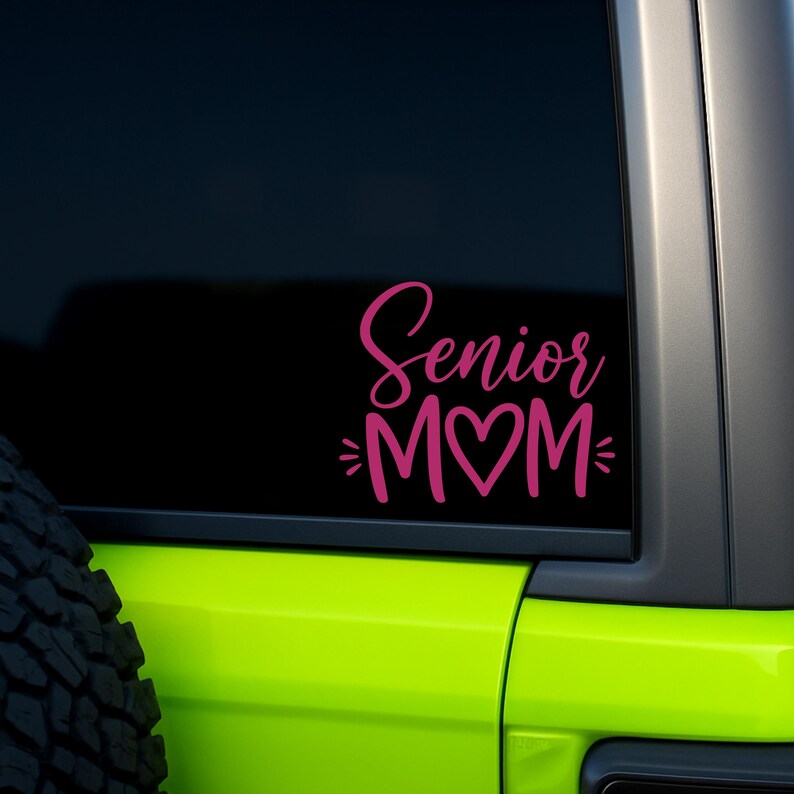 May include: A pink vinyl decal applied to a dark vehicle window. The decal features the text Senior in a cursive script font above the word MOM, where the letter O is replaced by a heart shape, flanked by two small horizontal lines on each side.