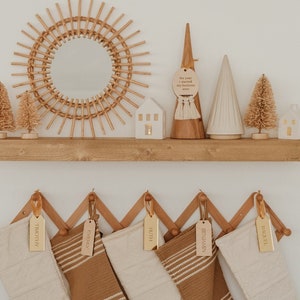 Op de afbeelding: Een houten wandplank toont een zonnespiegel, twee witte keramische huisjes, twee flessenborstelbomen en twee decoratieve kegels. Daaronder hangt een accordeonrek met vijf kousen voorzien van gepersonaliseerde gouden naamplaatjes.