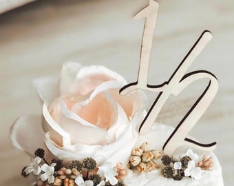 May include: A white cake with a single white rose and small white flowers on top. The cake is decorated with a wooden number "12" topper.
