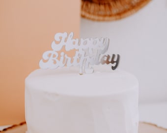 May include: A white cake with a silver "Happy Birthday" topper on top. The cake is on a wooden cake stand with a tan background.