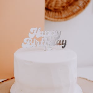 May include: A white cake with a silver "Happy Birthday" topper on top. The cake is on a wooden cake stand with a tan background.