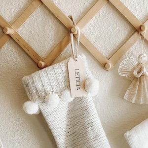 Op de afbeelding: Een uitschuifbaar houten wandrek met drie gebreide kerstsokken en een engelornament. De sokken zijn voorzien van gepersonaliseerde houten labels met de namen Zachary, Leticia en Victoria, waarbij de middelste sok is versierd met witte pompons.