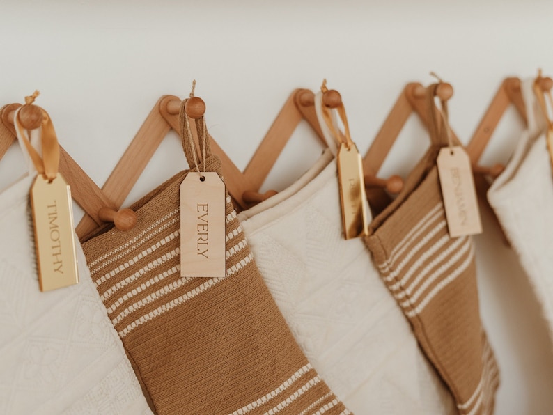Op de afbeelding: Een reeks hangende kerstsokken in afwisselende cr&egrave;me en beige kleuren met gestructureerde patronen, elk voorzien van een gepersonaliseerd rechthoekig naamplaatje van hout of goudkleurig materiaal, bevestigd met een lint aan een houten harmonica-rek.